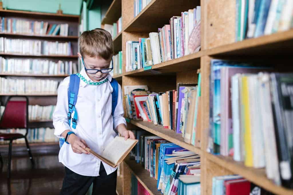 A young boy with glasses reads a book in a school library wearing a backpack.