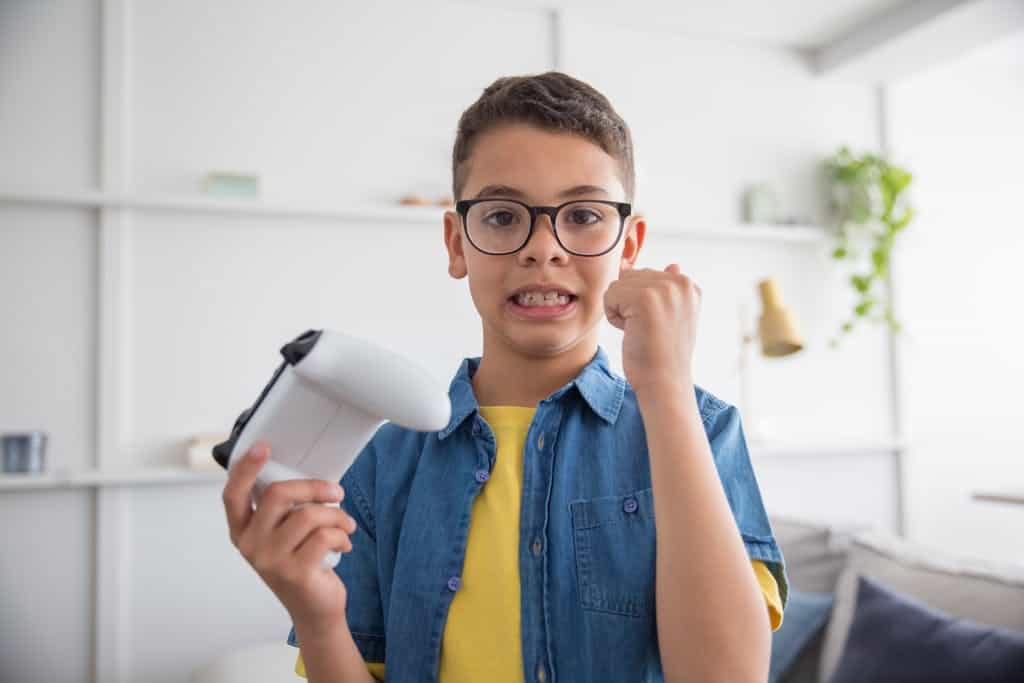 A young boy with eyeglasses eagerly plays video games indoors, holding a game controller. He's pumped about all of the elementary graduation gifts he received, but especially this one of a video game store gift card.