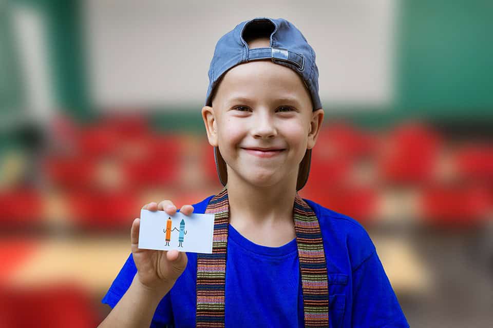 A 12 year old boy wearing a hat backwards ad a blue shirt is holding up a rebus puzzle card.