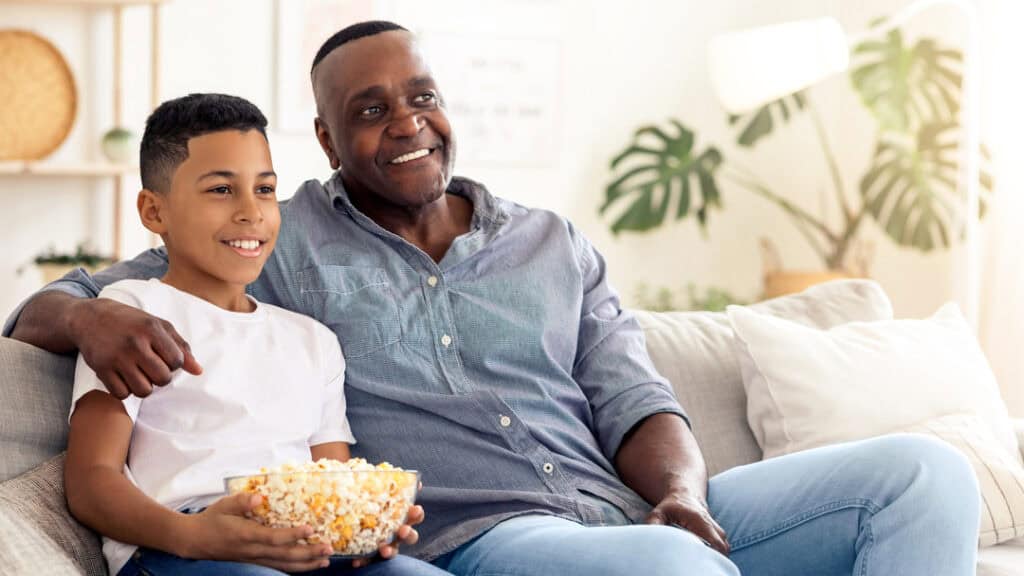 A grandfather and grandson share a cozy moment on the couch, enjoying a film together. They smile while holding a bowl of popcorn, creating cherished memories in their living room.