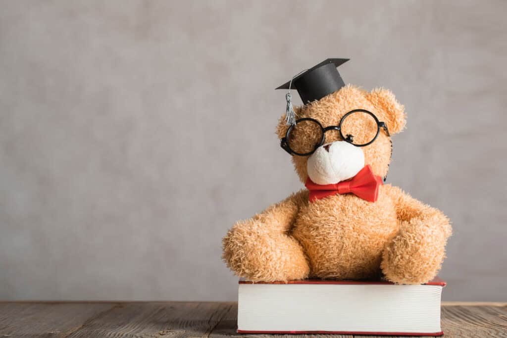 Brown teddy bear in an academic cap is placed on a thick dictionary. Closeup