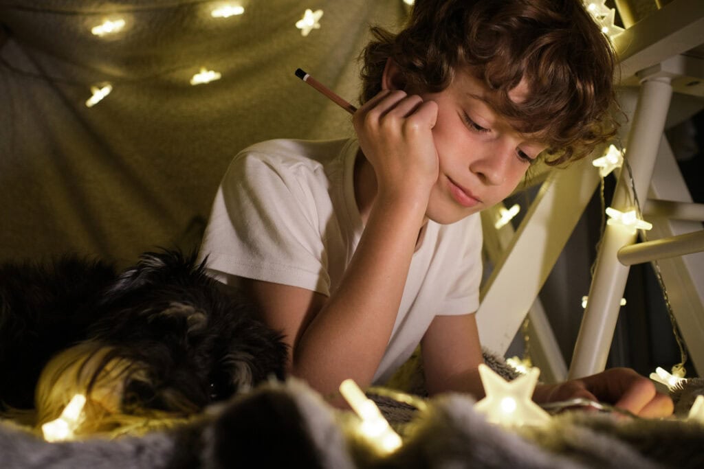 Focused boy lying under blanket fort with fairy lights glowing while spending time in cozy dark room at home. String lights are one of my favorite elementary graduation gifts for boys and girls.