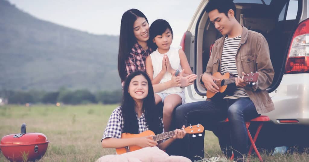 Happy teen playing ukulele with her family sitting in the car. They're enjoying a family road trip to celebrate their graduation.