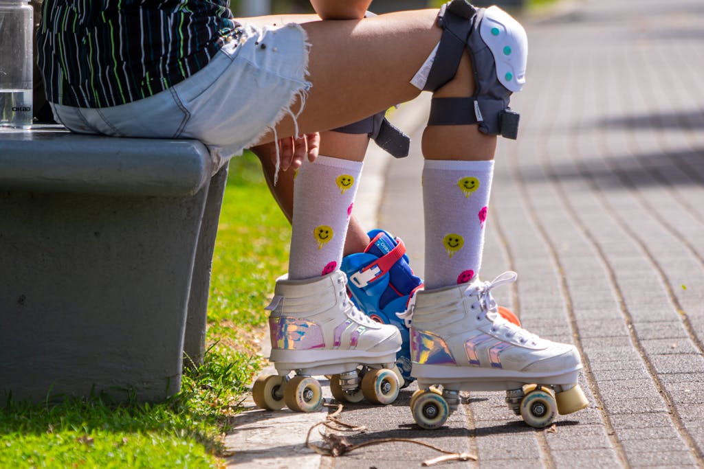 Young person wearing roller skates and knee pads sitting on a bench outside on a sunny day.