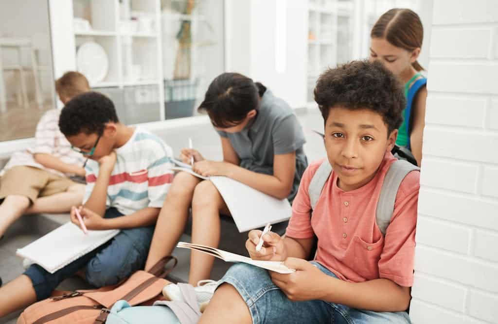 A diverse group of children sitting and studying together indoors with notebooks and backpacks.