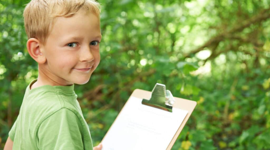 A young boy in a green shirt outside holding a clipboard. He is about to start an alphabet scavenger hunt.