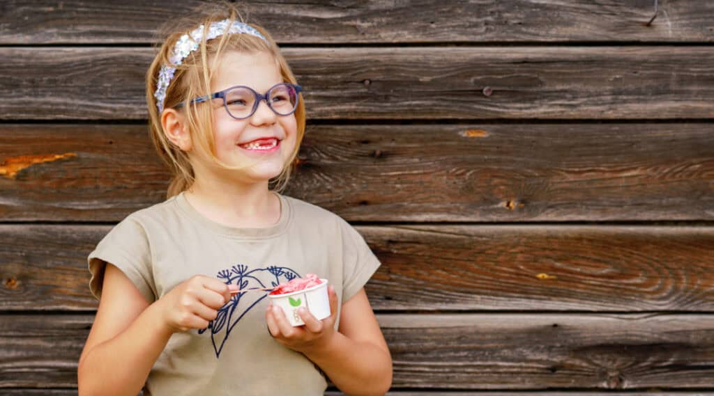 A 7-year old girl with glasses in a t-shirt smiling holding a bowl of ice cream.