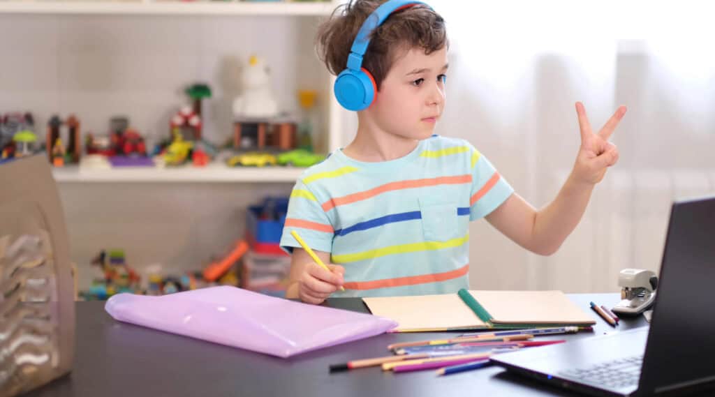 A young boy sitting at a desk wearing blue headphones. He's holding up two fingers to his lap top screen. He loves taking virtual summer camps.