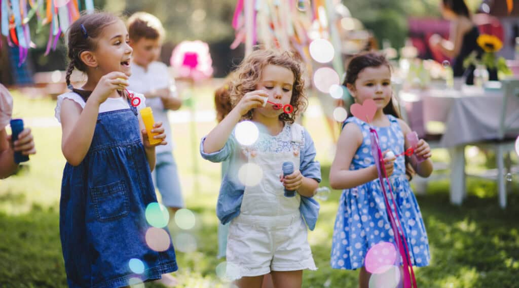 3 young girls are blowing bubbles outside in a backyard at a party that was planned and budgeted by kids.