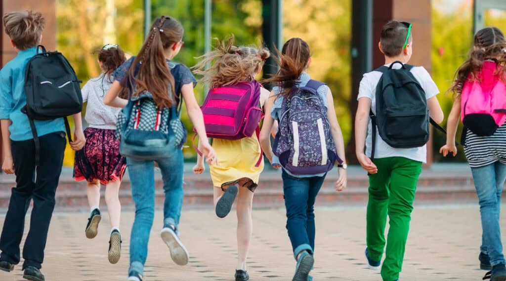 A group of boys and girls running into school pictured from behind so you can see all of their backpacks.