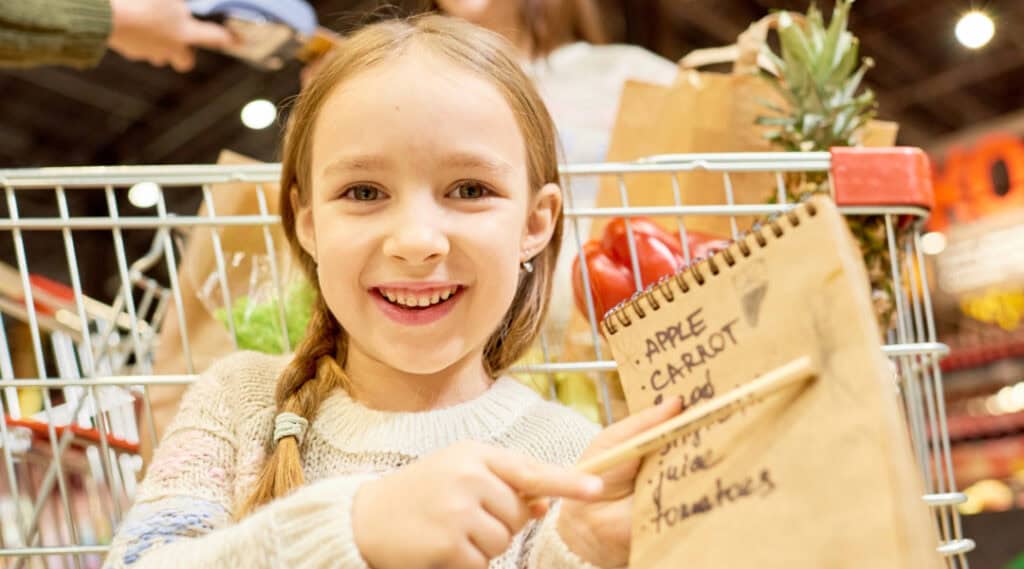 A young girl in a grocery store holding a grocery list to the camera.