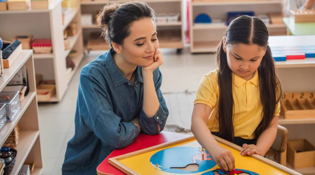 A teacher observing her student in a Montessori elementary classroom as the child works with maps.