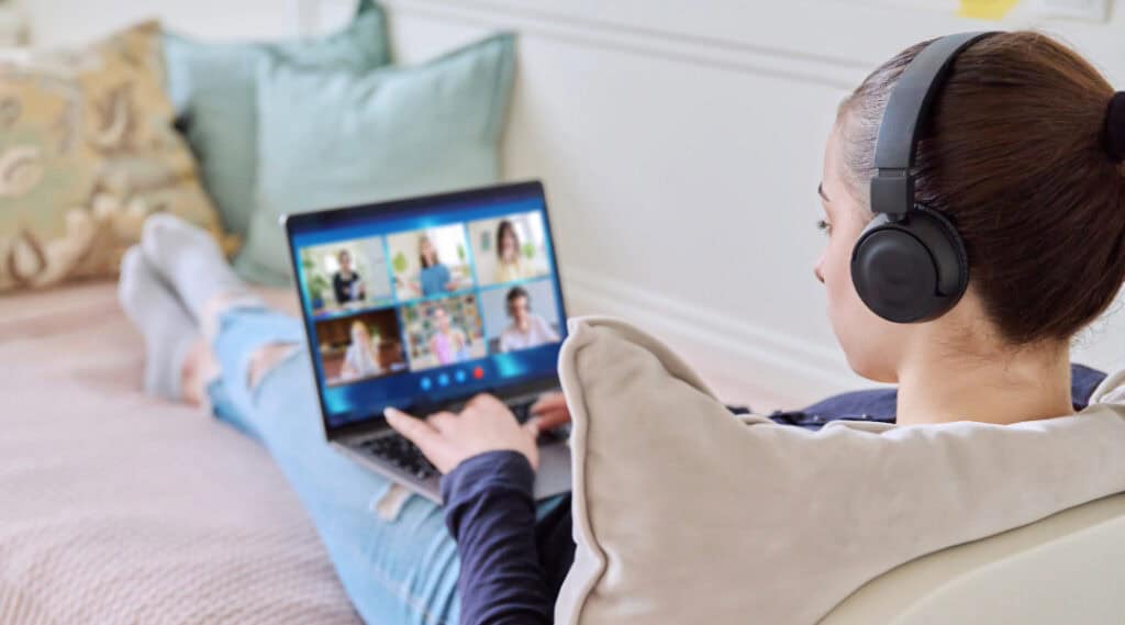 A teenage girl sitting on the couch with her feet up. She's taking a virtual summer camp on her iPad with a group of students online.