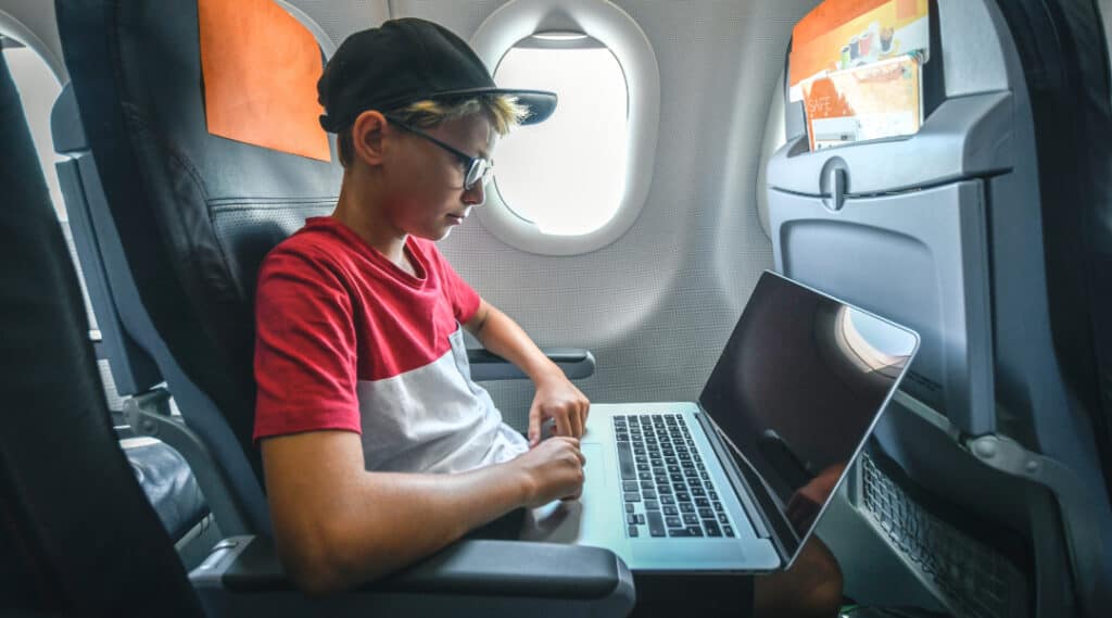 A young boy in a hat and glasses is sitting on an airplane with his computer on his lap.