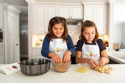Two young girls wearing Raddish aprons. They are getting into one of their subscription boxes for kids and going to learn how to cook.