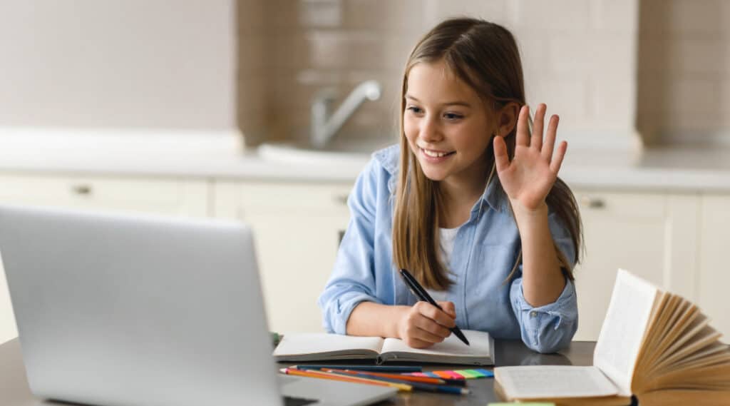 A young girl sitting at a desk looking and waving hello to her laptop. She is reaping the benefits of Outschool by taking online classes on the platform.