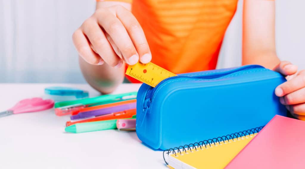A student in an orange t-shirt has one hand on and one hand placing a ruler in a blue pencil case. This is one of those must-have back to school supplies.