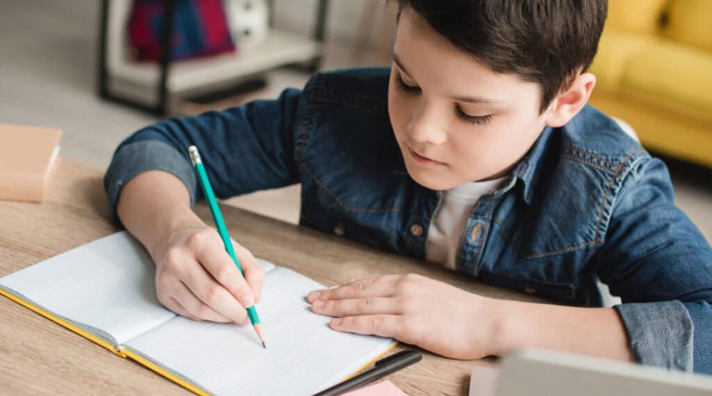 A young boy sitting at a table writing in a notebook.
