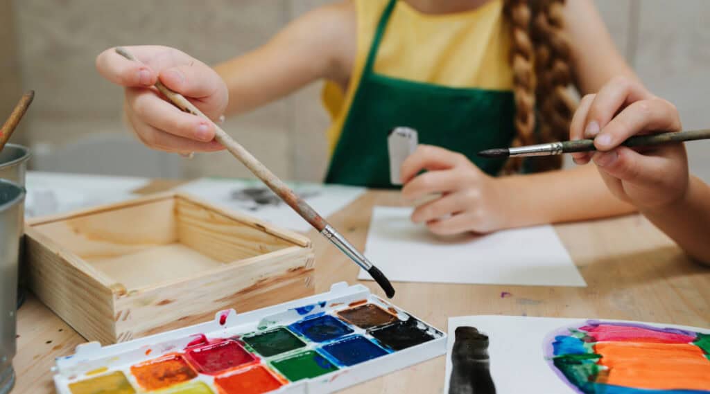 The hands of a young girl in a painters smock are shown reaching to dip her paintbrush into watercolor paint. 