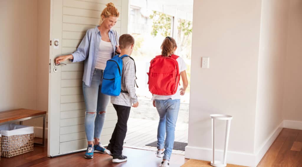 An image of a mom holding the door open while her two children walk out on their way to school. One of my fav back to school tips for families is to have a basket at the front door for the kids school items to go in. 