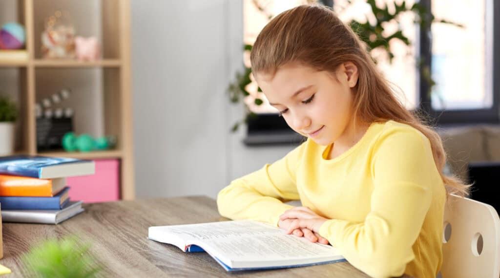 A young girl sitting at a table at home reading a book. She's taking some time before going to school to quietly read.