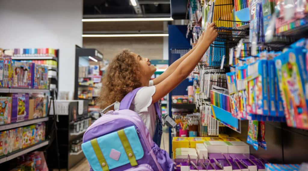 A young girl wearing a purple back pack reaching for a notebook in the store. She is back to school shopping.