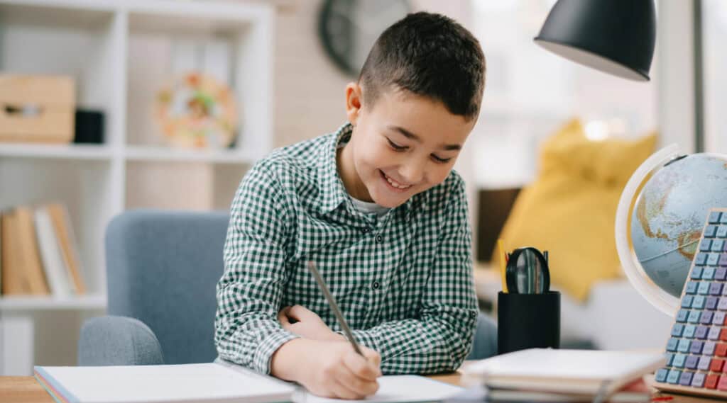 A 10-year-old boy sitting at a table at home doing homework—a great example of one of our favorite back to school tips for families.