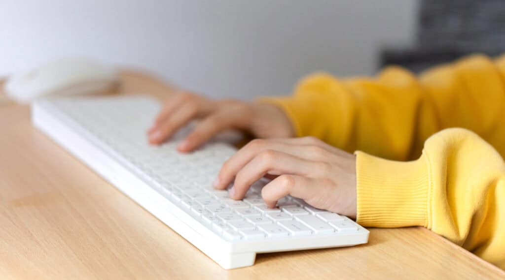 A set of child's hands in a yellow sweatshirt typing on a wireless keyboard.