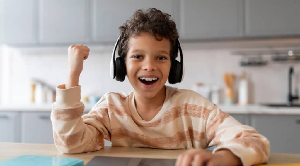 A young boy sitting at a table with wireless headphones with a big smile and fist pump. He is excited about his back to school supplies.