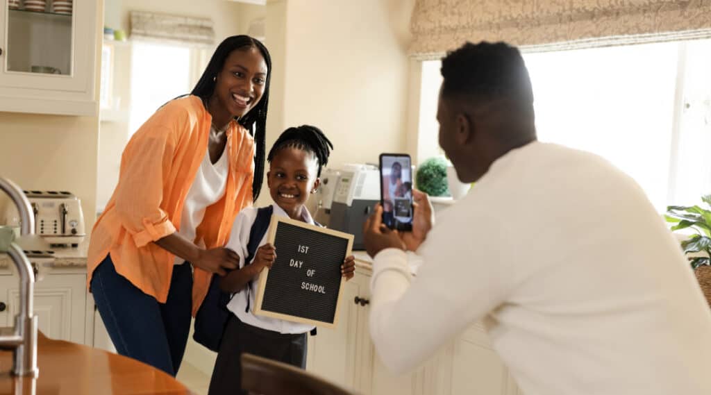 A mom and daughter pose for a picture while dad takes the photo with his phone. The girl is holding a sign that says 'back to school'