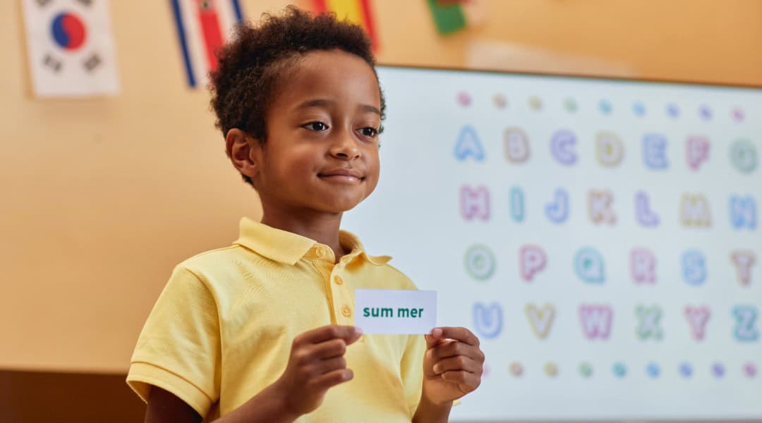 A grade 2 student in a yellow shirt standing holding a card that has the word 'summer' on it. He is going to sort it into the correct part of speech.