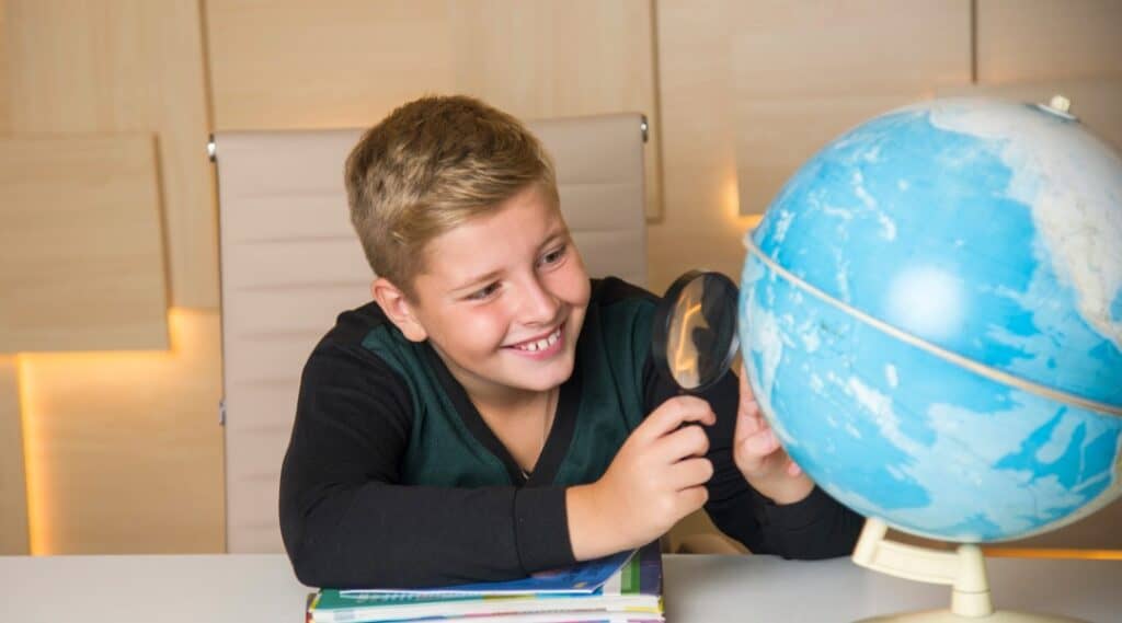 A picture of a 10 year old boy sitting at a desk looking at a globe with a magnifying glass. He is happy with his Little Passports subscription box.