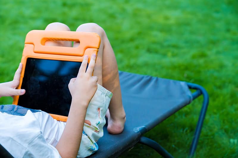 A young child is lounging on a sunbed in a backyard, holding a tablet in an orange case. The shot captures a moment of digital relaxation in nature, with a vibrant green lawn in the background.