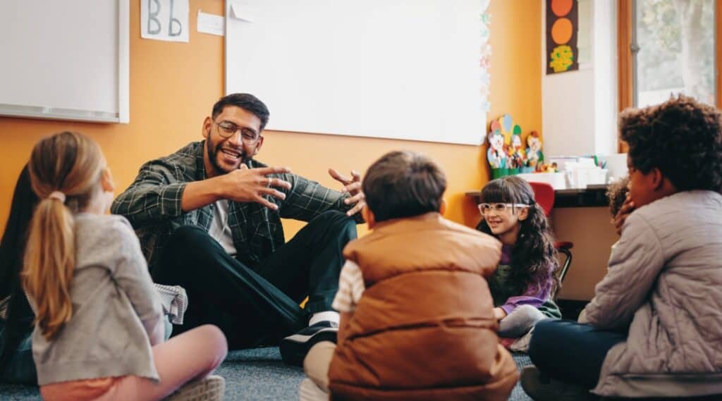 AN image of a male teacher sitting on the floor with students seated in a circle. They are discussing one of Tony Robbins quotes for kids.