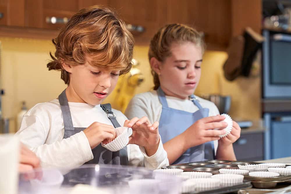 Focused boy and girl preparing baking paper in metal pan for homemade cupcakes while helping crop parent with housework in kitchen