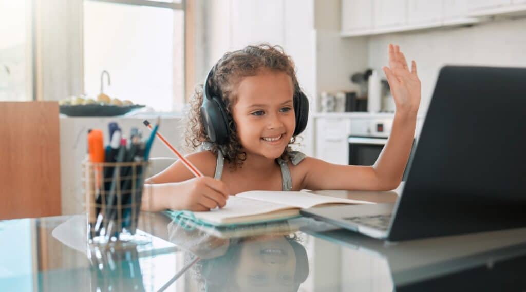 A young girl standing in her kitchen with headphones on and a lap top. She's taking an online class through Outschool.