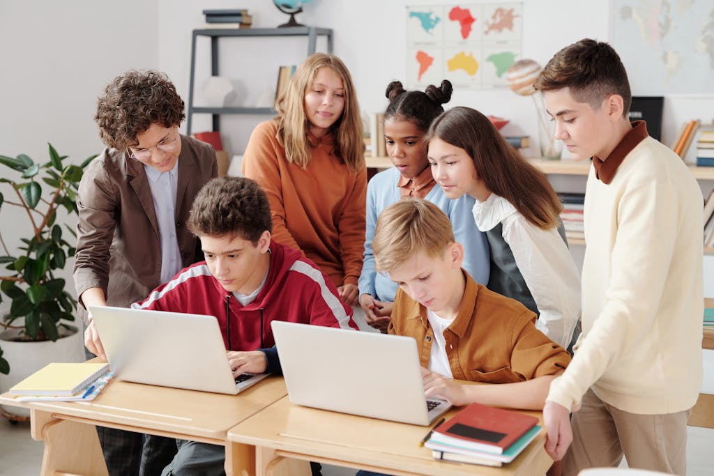A group of diverse students watching educational videos on laptops in a classroom setting, guided by a teacher who breaks into a teachable moment.