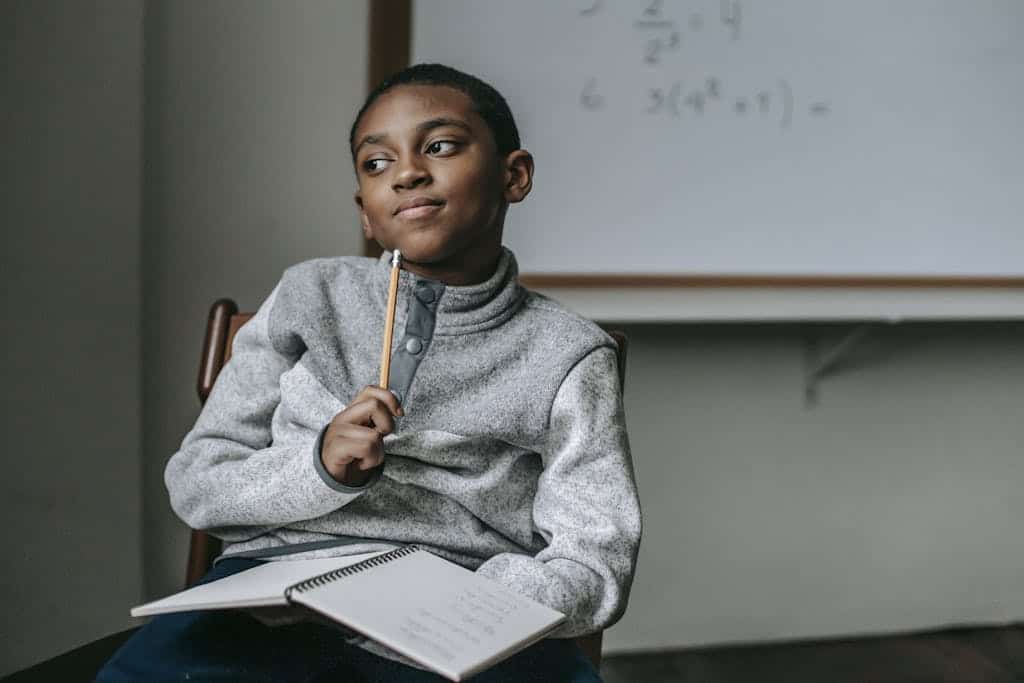Concentrated African American boy in casual outfit sitting on chair with pencil and copybook and thinking about his time management skills.
