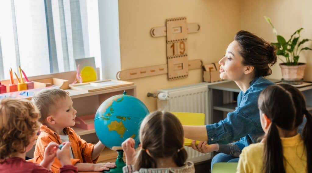 A teacher and young elementary students sit down for a lesson. In order to understand this pedagogy, you need to know who Maria Montessori is.