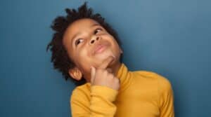 A 6 year old boy wearing a yellow shirt with his hand on his chin while he looks up thinking. He's wondering what Montessori is.