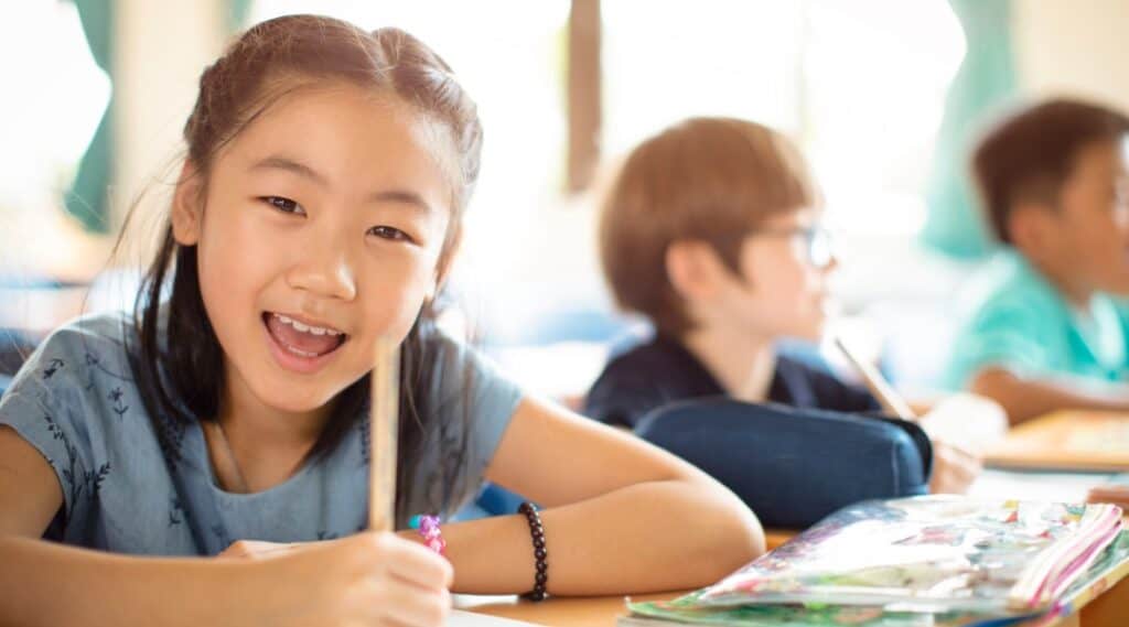 A young elementary student smiling, sitting at a table holding a pencil working on learning the word of the week.