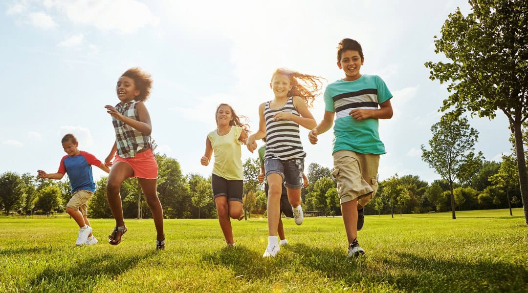 A picture of 5 children running toward the camera in an open green space. It's a sunny summer day.