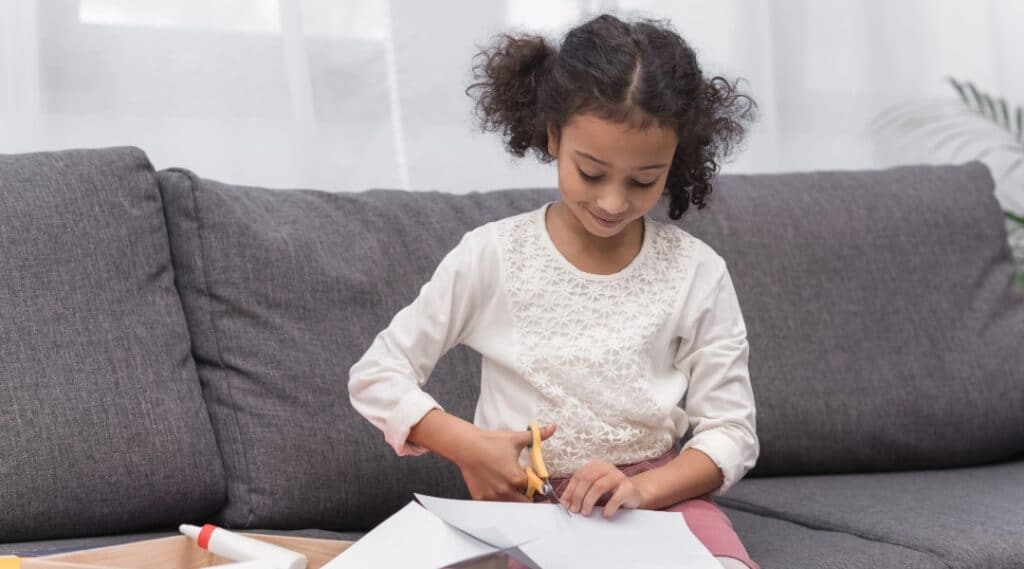 A young girl is sitting on a couch cuttingout her movement dice 2D net so she can fold it into a cube and play Just Roll With It.