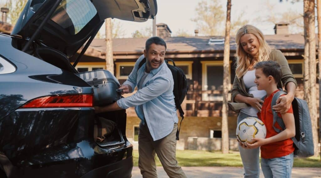 A dad packs the trunk of the car while mom and son watch and smile. They are ready for some road trip games!